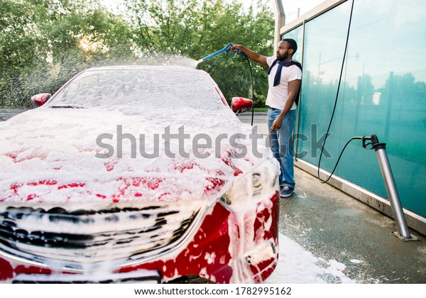 Young black man cleaning his\
red luxury car with foam spray outside at self car wash station.\
Casual African guy washing his car with high pressure sprayer and\
foam