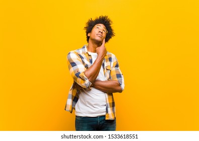 Young Black Man With A Concentrated Look, Wondering With A Doubtful Expression, Looking Up And To The Side Against Orange Wall