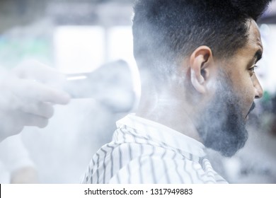 Young Black Man Customer Received New Haircut In Barbershop.Professional Barber Pour Hoist On Clients Neck Before Shaving With Sharp Razor In Male Beauty Salon.Portrait Of African Boy In Barbers Chair