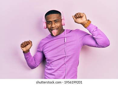 Young Black Man Dancing Listening To Music Using Headphones Sticking Tongue Out Happy With Funny Expression. 