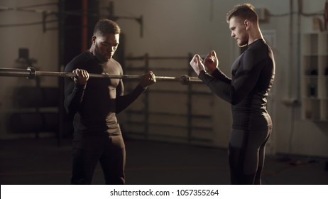 Young Black Man Doing Exercises With A Personal Trainer As He Lifts A Barbell Weight To Chest Height In A Gym In Evening