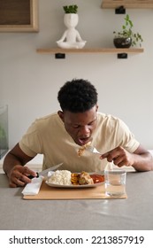 Young Black Man Eating Fried Chicken, Rice And Plantain With Tomato Sauce Plate In The Living Room.