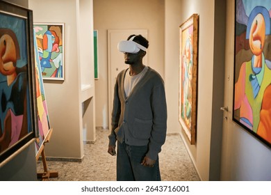 Young Black man exploring virtual reality art exhibition while wearing headset in modern art gallery surrounded by vibrant abstract paintings