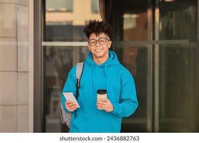 Young black man with eyeglasses takes coffee break while leisurely browsing online on his smartphone in urban setting, smiling to camera. Happy student guy standing outside college building - Powered by Shutterstock