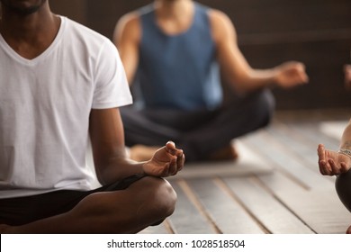 Young black man and a group of young sporty people practicing yoga lesson, sitting in Sukhasana exercise, Easy Seat pose, working out, indoor close up focus on mudra gesture, studio room