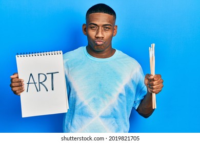 Young Black Man Holding Art Notebook And Painter Brushes Looking At The Camera Blowing A Kiss Being Lovely And Sexy. Love Expression. 