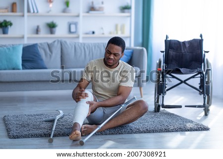 Young black man with injured leg sitting on floor after falling down, having trouble walking with crutches, wheelchair standing nearby. African American guy using mobility aid with no success