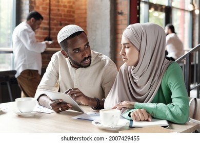 Young Black Man In Kufi Cap Using Tablet While Explaining Data To Female Colleague In Coffee Shop