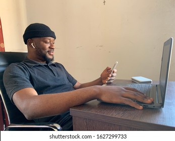 Young black man listening to songs on his wireless earphones and pressing his phone while using his laptop