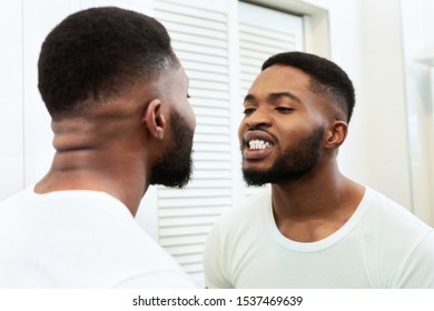 Young Black Man Looking In Mirror At His Mouth And Teeth In Bathroom, Checking Their Health State