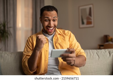 Young Black Man Looking At Smartphone In Excitement, Making YES Gesture At Home. Emotional Millennial Guy Winning Lottery Or Casino Bet Online, Gambling Or Playing Video Game Indoors