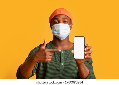 Young Black Man In Protective Medical Mask On Face Pointing At Smartphone With Blank White Screen, Millenial African Guy Showing App With Coronavirus Outbreak Tracking Map, Yellow Background, Mockup