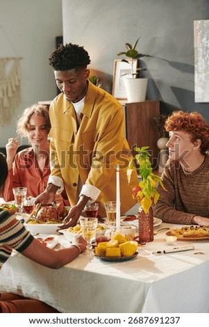 Young Black man serving roast chicken to diverse group of young adults sitting around dining table sharing meal, smiling and interacting during Thanksgiving gathering, visible food and drinks