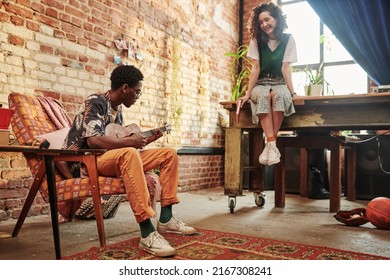 Young Black Man Sitting In Armchair And Playing Guitar For His Girlfriend Relaxing On Table In Front Of Him In Living Room Of Loft Apartment