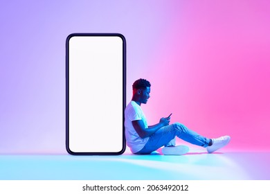 Young Black Man Sitting Next To Huge Smartphone With Empty Screen, Advertising New Mobile Application Or Website, Using Modern Gadget In Neon Light. Cellphone Display Mockup