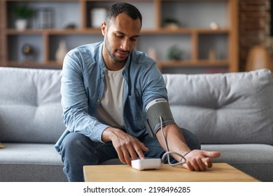 Young Black Man Sitting On Couch And Checking Blood Pressure With Upper Arm Monitor, Millennial African American Guy Measuring Arterial Tension While Relaxing In Living Room At Home, Copy Space