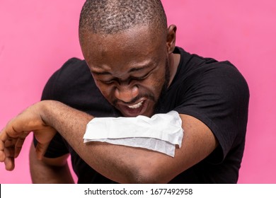 Young Black Man Sneezing Into A Napkin On His Elbows