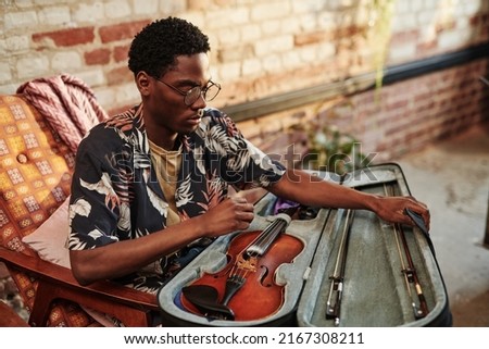 Young black man in stylish casualwear opening slipcover with violin and fiddlesticks while sitting in armchair in living room of loft apartment
