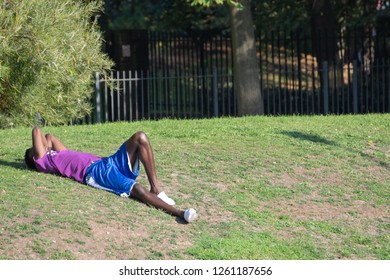 A Young Black Man Sunbathing On Grass In A London Park Wearing Sports Clothes (white Trainers/sneakers, A Purple T-shirt And Blue Shirts). He Is Covering His Face With His Arms To Block Out The Sun