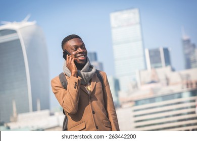 Young Black Man Talking On Mobile Phone In London With City Skyscrapers On Background In A Sunny Day. He Is Wearing A Coat And Has A Vintage Backpack.