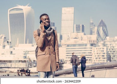 Young Black Man Talking On Mobile Phone In London With City Skyscrapers On Background In A Sunny Day. He Is Wearing A Coat And Has A Vintage Backpack. Vintage Filter Applied.