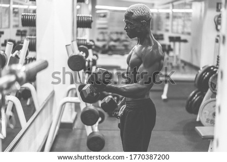 Young black man training with dumbbells inside gym during night time - African guy doing fitenss workout session - Sport and body building concept - Focus on face - Black and white editing
