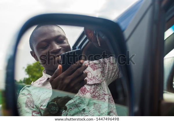 young black man using his phone while\
standing next to his car, seen from the side\
mirror