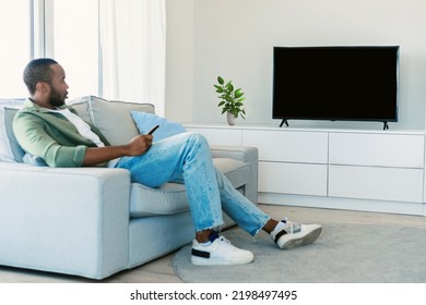 Young Black Man Watching Tv With Empty Screen, Resting On Sofa In Living Room Interior, Mockup. Entertainment With Modern Technology At Spare Time, News And New Movie