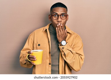 Young black man wearing glasses and drinking a cup of coffee covering mouth with hand, shocked and afraid for mistake. surprised expression  - Powered by Shutterstock