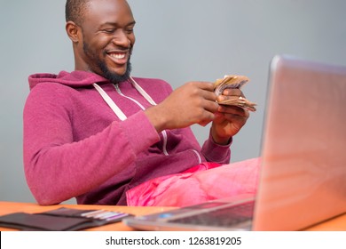 Young Black Man Wearing A Hoodie Smiling And Counting Money With A Laptop And A Wallet On His Desk