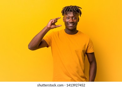 Young black man wearing rastas over yellow background holding something little with forefingers, smiling and confident. - Shutterstock ID 1413927452
