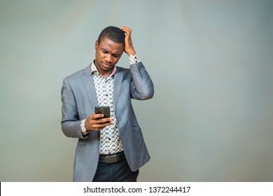 Young Black Man Wearing A Suit Holding His Phone And Scratching His Head Worried And Confused About Something