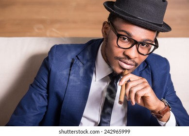 Young Black Man Is Wearing Suit And Hat, Smoking A Cigar.