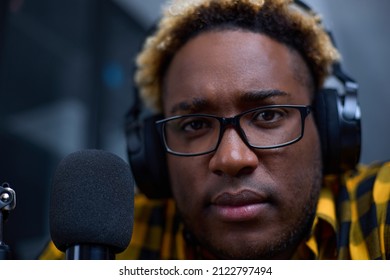 A Young Black Man Wearing Wireless Headphones Broadcasts His Podcast Over The Internet Using A Professional Microphone In A Small Broadcasting Studio. Close-up Of A Blogger Or Streamer Wearing Glasses