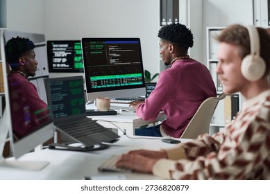 Young Black man working at computer with code on screen in office, sitting next to young man wearing headphones, multiple monitors displaying programming interface