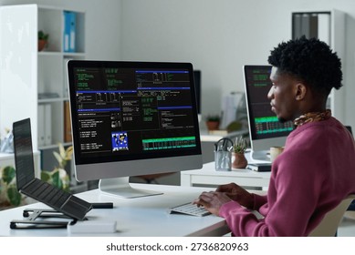 Young Black man working at computer with multiple monitors displaying programming code in office, focusing on software development tasks, hands on keyboard, sitting at desk