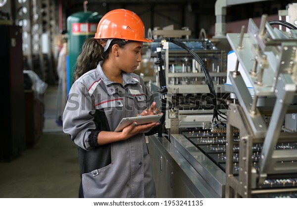 A young black manufacturing worker\
controls an assembly line in a factory, making notes on a tablet.\
Young woman in hard hat watches the work\
process