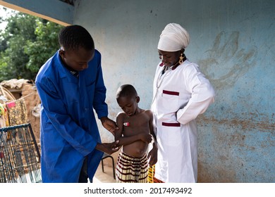 Young Black Medical Assistenst Is Taking The Temperature Of A Small Toddler During A Home Visit, Under The Close Supervision Of A Female Pediatrician During Health Screening Of Children In A Village