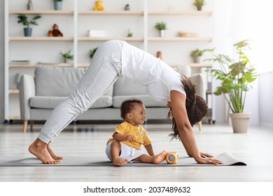 Young Black Mother Practicing Yoga At Home With Her Infant Son, Standing In Dog Pose While Training On Fitness Mat In Living Room, Making Back Stretching Exercise And Smiling To Her Baby, Copy Space