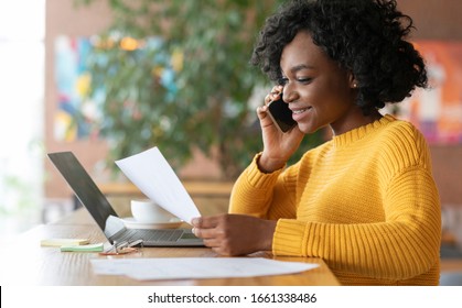 Young black smiling woman editor talking to clients on phone, working with laptop at cafe