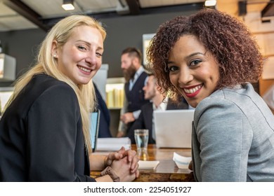 Young black south american woman looking at camera with her blonde caucasian colleague while working in modern office - Break from work and coworking concept