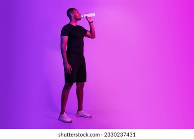 Young black sportsman in sportswear drinking water on purple studio background, posing with fitness bottle, standing near free space. Full length of sporty guy staying hydrated after workout - Powered by Shutterstock