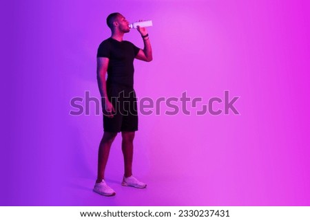 Young black sportsman in sportswear drinking water on purple studio background, posing with fitness bottle, standing near free space. Full length of sporty guy staying hydrated after workout