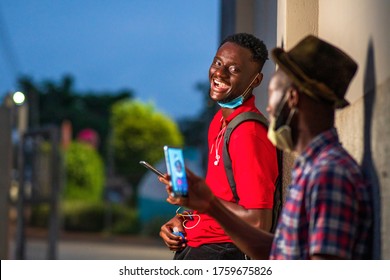 Young Black Student Back To Campus ,laughing And Wearing Surgical Face Mask,holding Phone In Hand, Talking To A Friend Blurred Out,making A Voice Or Video Call,sharing Social Media Content,conversing