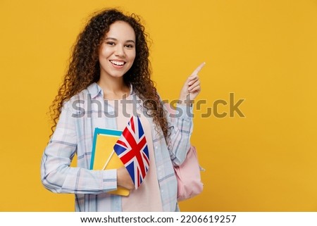 Young black teen girl student she wear casual clothes backpack bag hold books british flag point finger aside on area isolated on plain yellow color background. High school university college concept