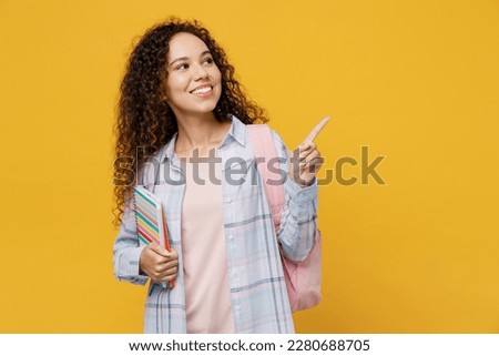 Young black teen girl student she wear casual clothes backpack bag hold books point index finger aside on workspace isolated on plain yellow color background. High school university college concept