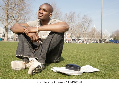Young black tourist man sitting down on green grass in the city of London while visiting, smiling.