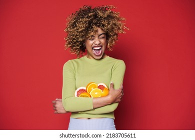 Young Black Woman With Afro Curls Having Fun Holding Oranges Cut In Half Isolated Over Red Background