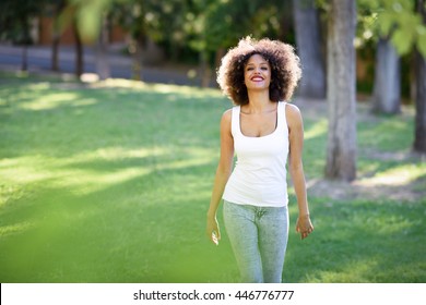 Young Black Woman With Afro Hairstyle Smiling In Urban Park. Mixed Girl Wearing White T-shirt And Blue Jeans Walking On The Grass.