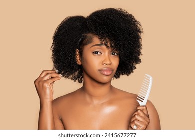 Young black woman appears sad as she touches her curly afro hair and holding white comb, standing against soft beige background - Powered by Shutterstock